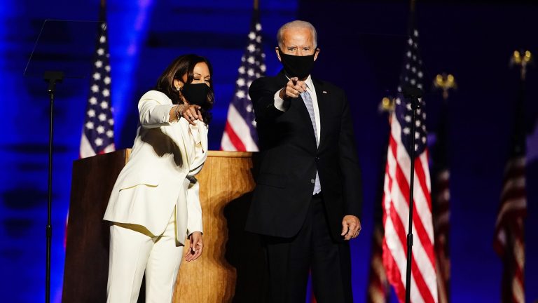 Joe Biden and Kamala Harris wearing masks, standing at a lectern pointing towards the audience.