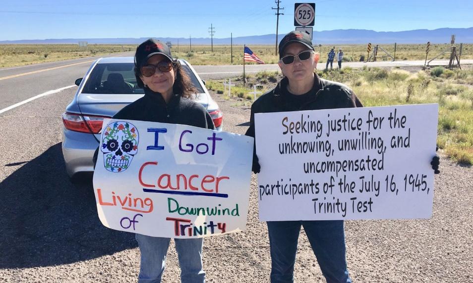 Two activists hold signs. One says "I got cancer living downwind of Trinity". The other says "Seeking justice for the unknowing unwilling and uncompensated participants of the July 16, 1945 Trinity Test".