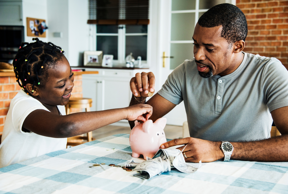 A smiling Black father and daughter put change into a piggy bank.
