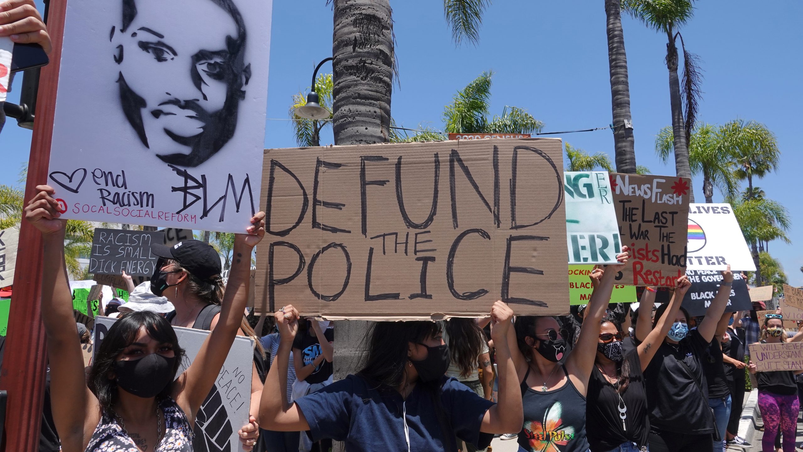 Photo of people at rally holding cardboard sign that says "DEFUND THE POLICE" with a white sign nearby showing a black outline stencil of MLK Jr. and "BLM" written on the bottom.