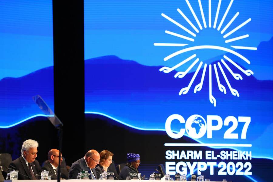 United Nations leaders seated at tables with microphones on stage at the COP27 climate conference in Egypt. There is a white logo that looks like a sun and the rays on the bottom look like outstretched hands. Beneath the logo reads "COP 27 Sharm El Sheikh Egypt 2022."