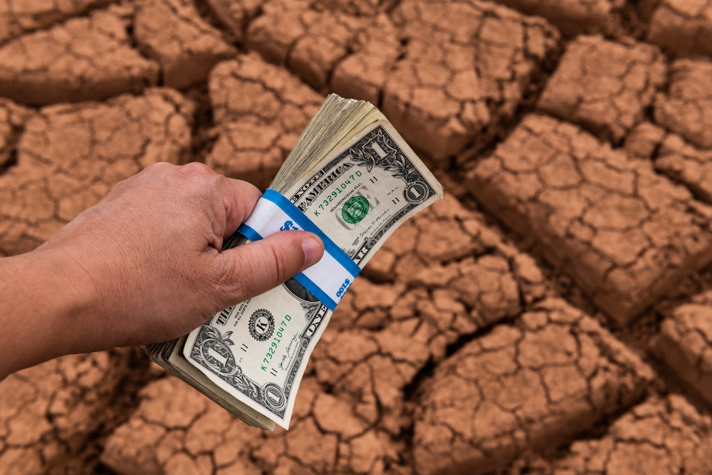 An aerial view of the left hand of a person holding a stack of $1 bills with cracked desert like earth beneath.