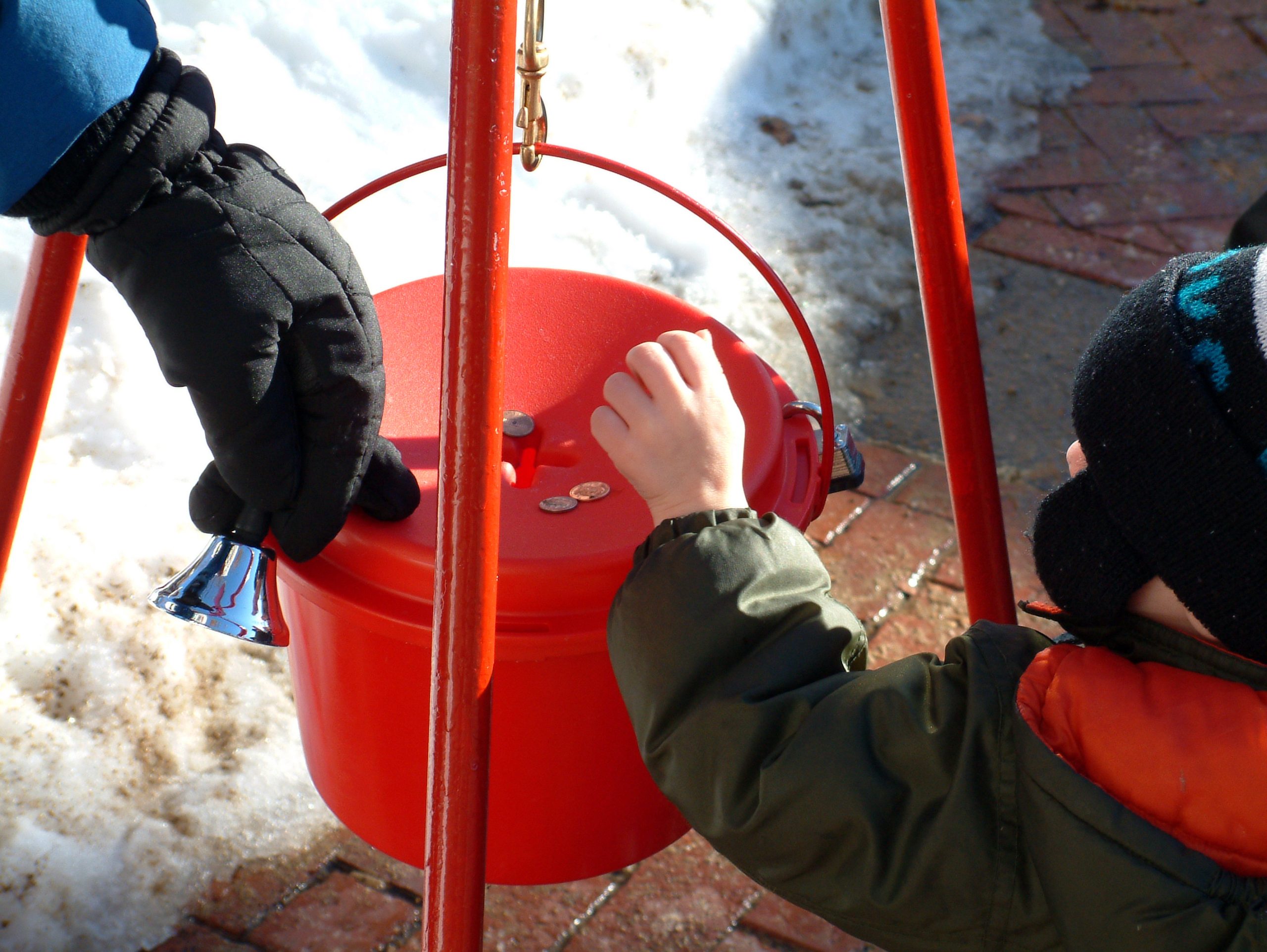 Image of a small child dressed in winter clothing, putting money in a red salvation army change kettle steadied by a gloved hand, and there is snow in the background of the photo.