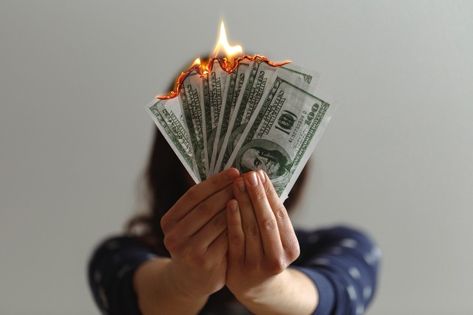 A person with long and a blue shirt has their arms outstretched with money fanned out in hands. The money is on fire and covering their face.