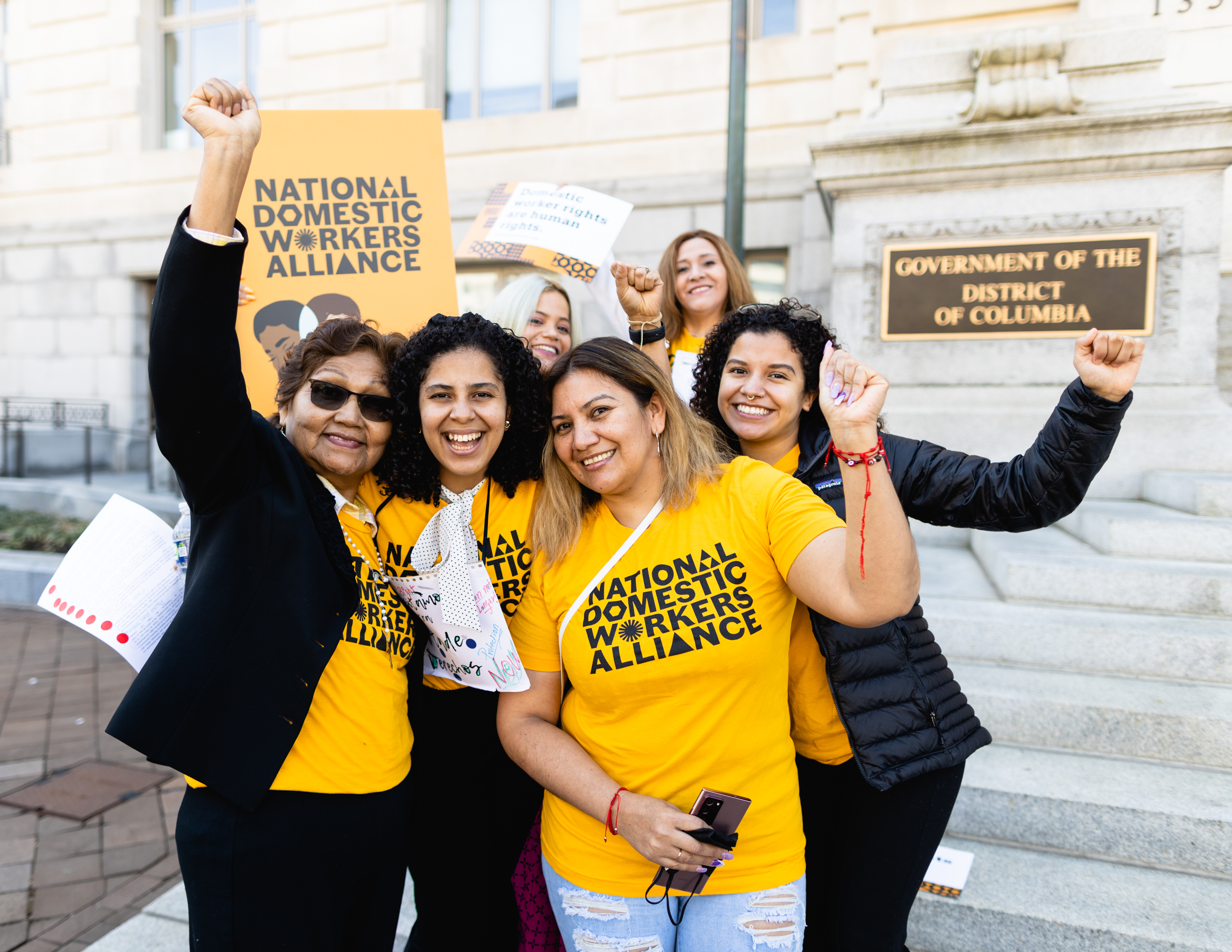 Member of the Domestic Workers Alliance wearing their yellow shirts, posing for a picture with fists raised in the hair. There is a sign in the background showing support of the Domestic Workers Bill of Rights.