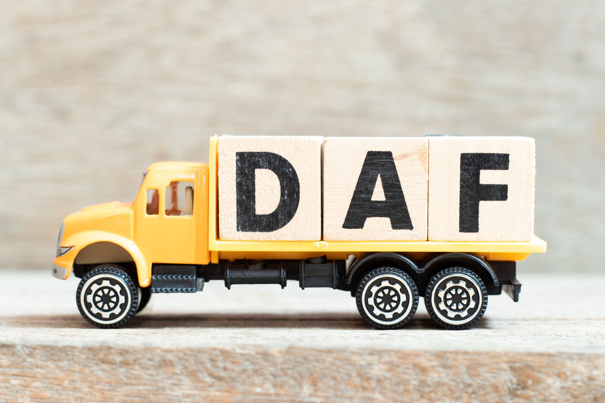 Profile view of yellow toy truck with flat bed carrying three wooden blocks that have "D" "A" "F" printed on them.