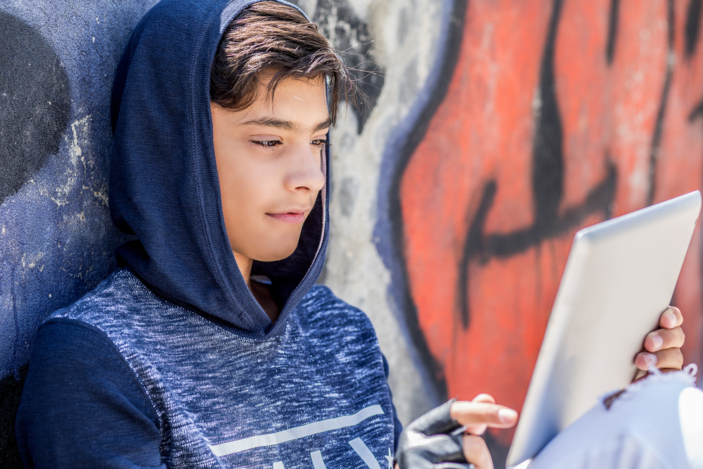 Young person with a blue hoodie on, seated on the ground, leaning against the wall, using a laptop.