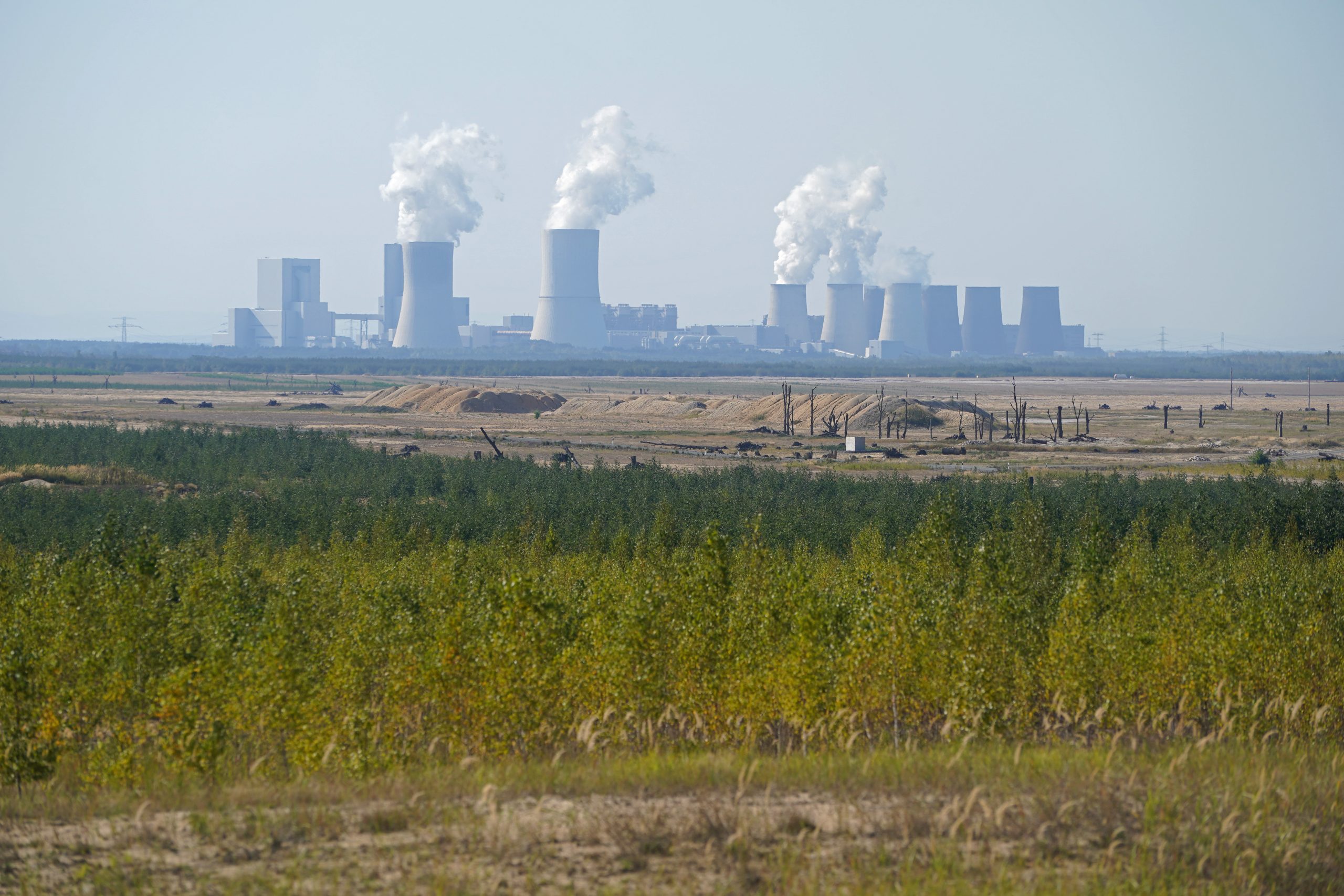 Image of coal plants far off in the distance emitting pollution, with green grass and flatlands in the foreground.