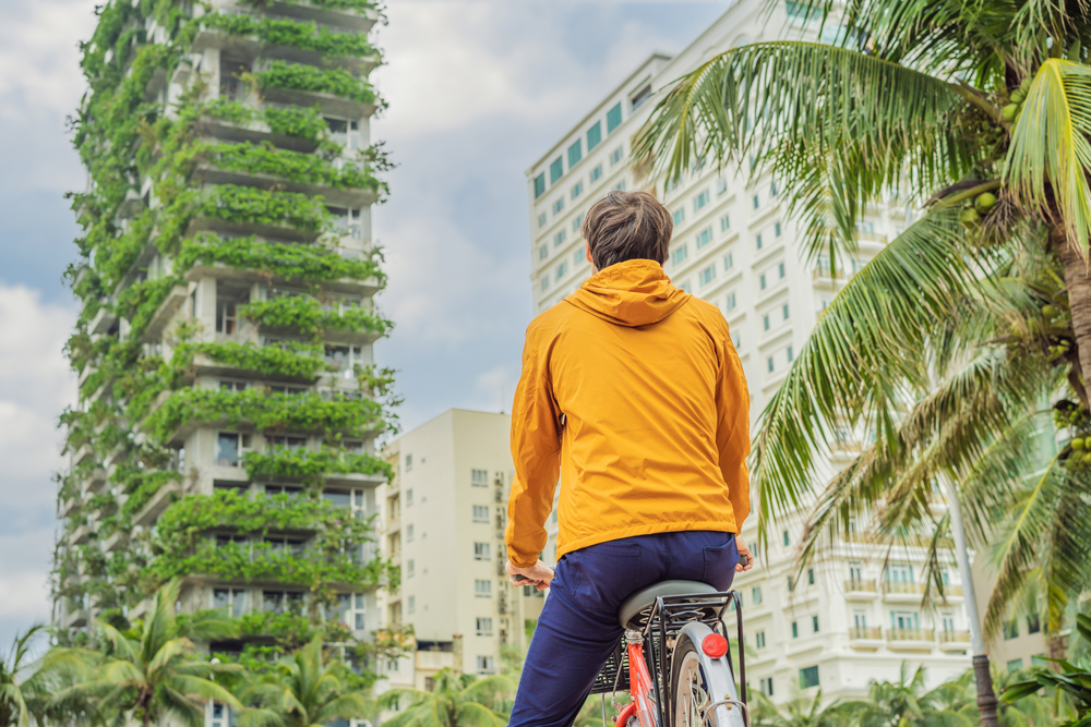 Shot of someone with their back to us in yellow jacket. They're on a bike and gazing upon a building with lush greenery growing on it.