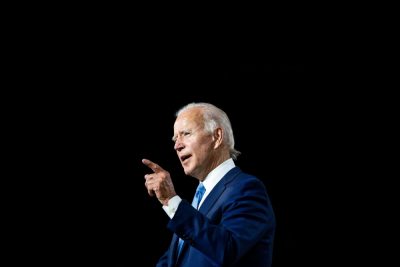 A profile shot of President Biden in a blue suit and white shirt, speaking and pointing his finger.